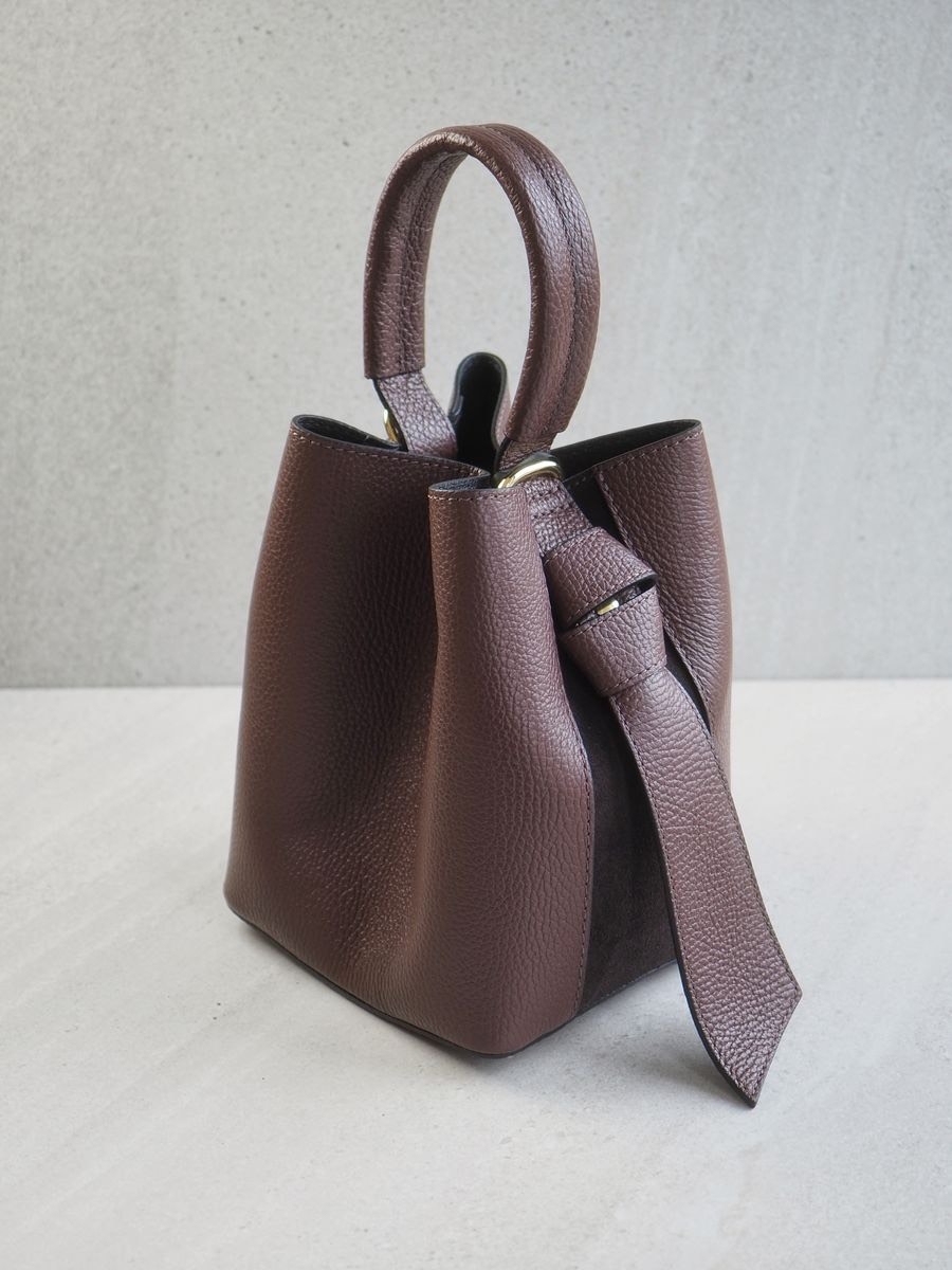 Sofistikert og elegant bøtteveske i både semsket og glatt skinn. Vesken har et dekorativt knutebelte. Vesken har to stropper, den lengste er regulerbar, den andre er et håndtak.